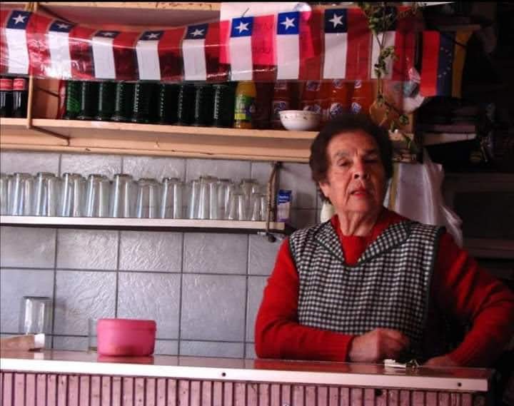 La tía Tita detrás del mesón de un local adornado con banderas chilenas