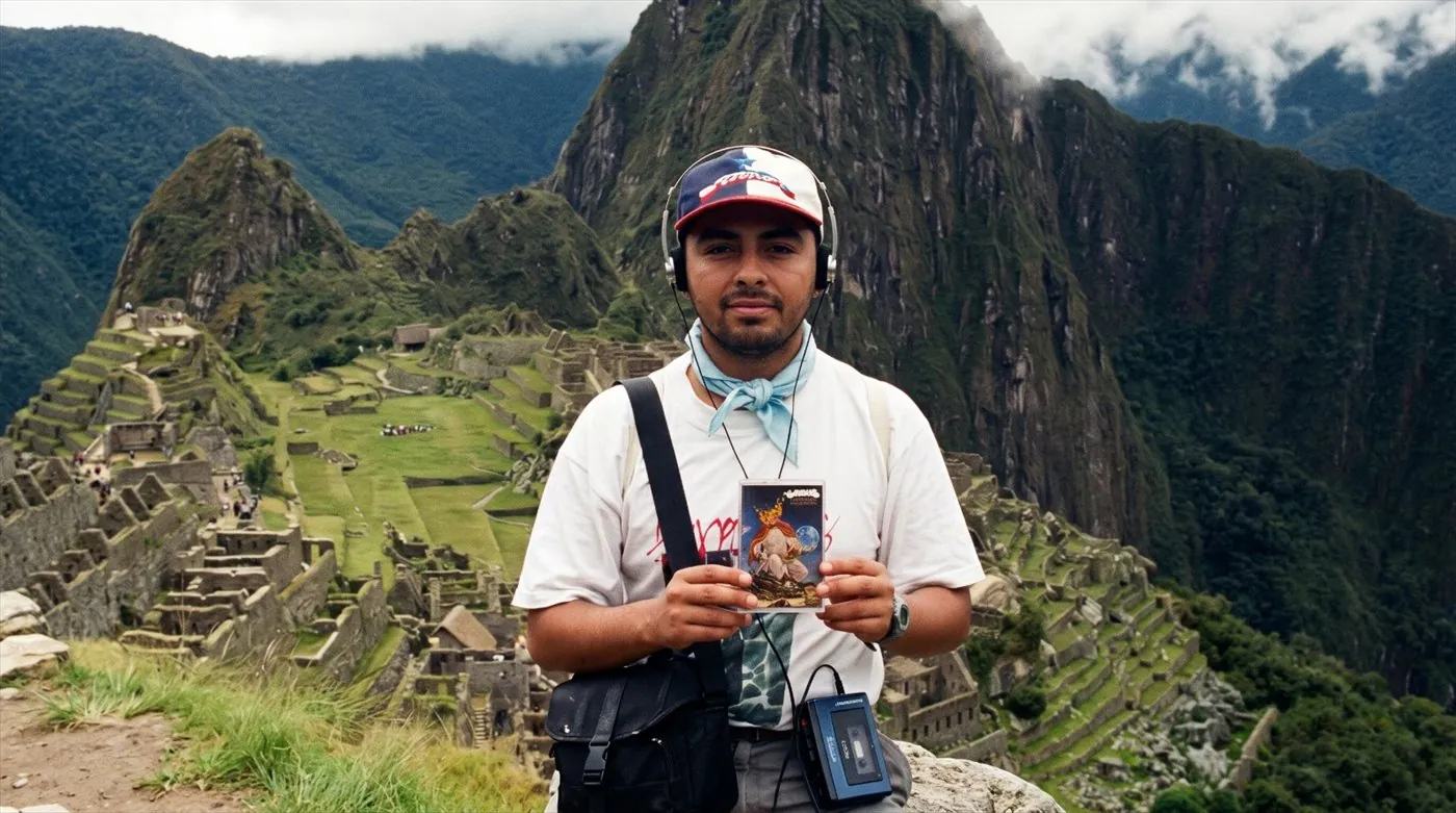 Tuga, con audífonos y un walkman, muestra el cassette de «Alturas de Machu Picchu» de Los Jaivas frente a las ruinas de Machu Picchu