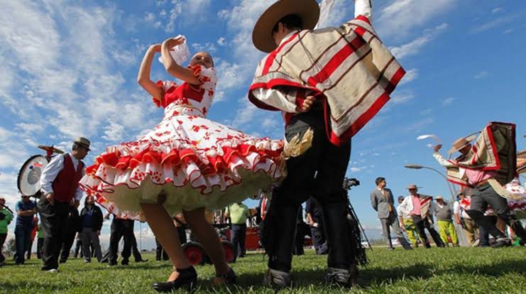 Pareja bailando cueca al aire libre, ella con vestido de huasa y él con manta y chupalla